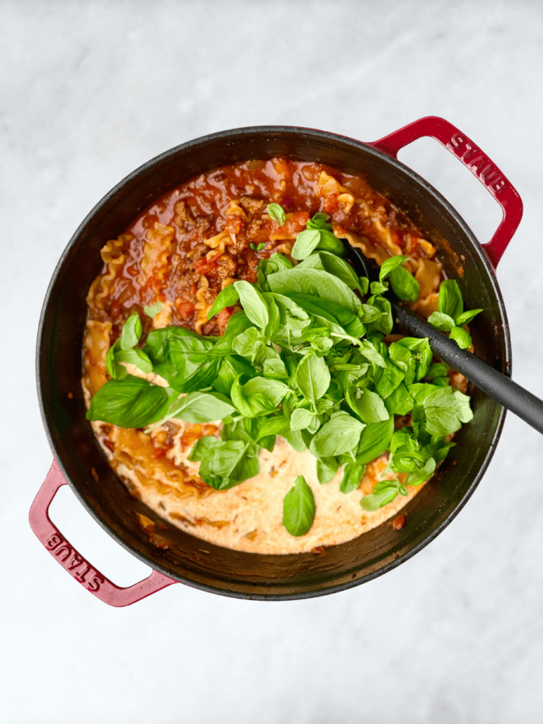 Overhead view of creamy high-protein lasagna soup simmering in a red Dutch oven with basil and heavy cream added.