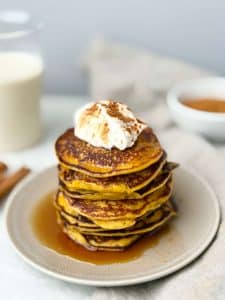 Close-up of golden pumpkin protein pancakes stacked with whipped topping and maple syrup drizzle.