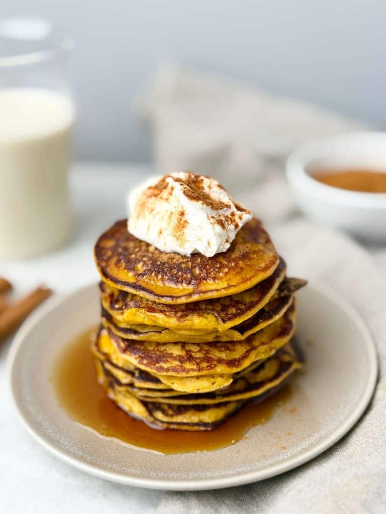 Close-up of golden pumpkin protein pancakes stacked with whipped topping and maple syrup drizzle.