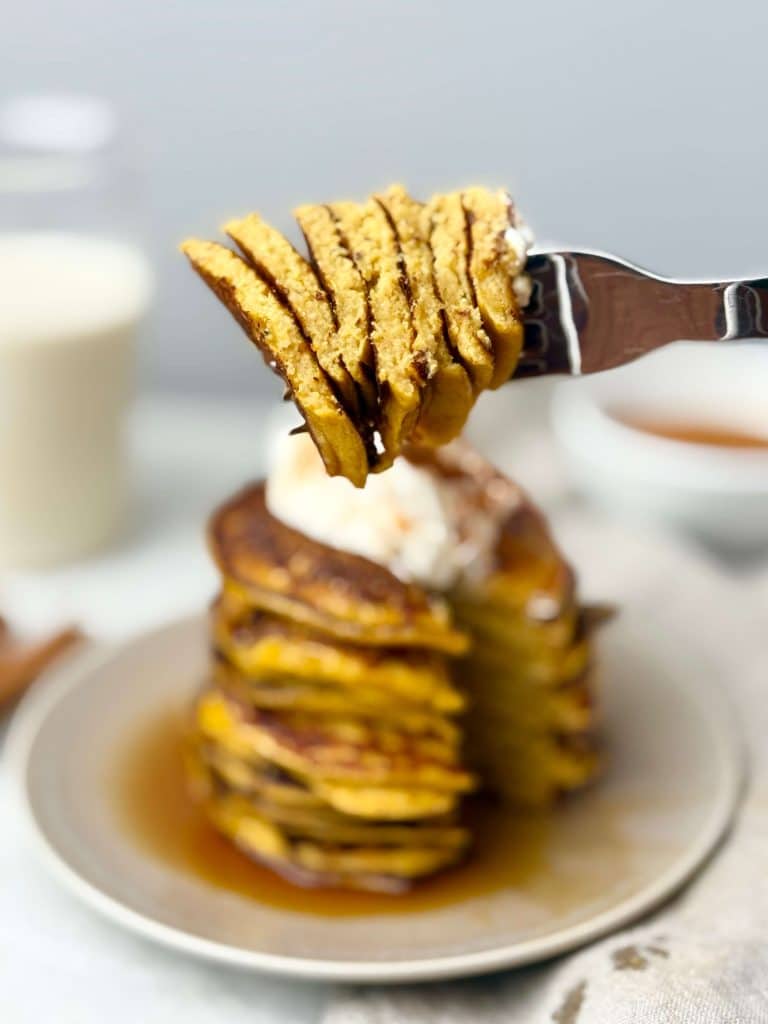 Fork lifting a bite of fluffy pumpkin protein pancakes over a syrup-drizzled stack on a beige plate.