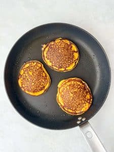 Three pumpkin spice protein pancakes cooking in a nonstick skillet until golden brown.
