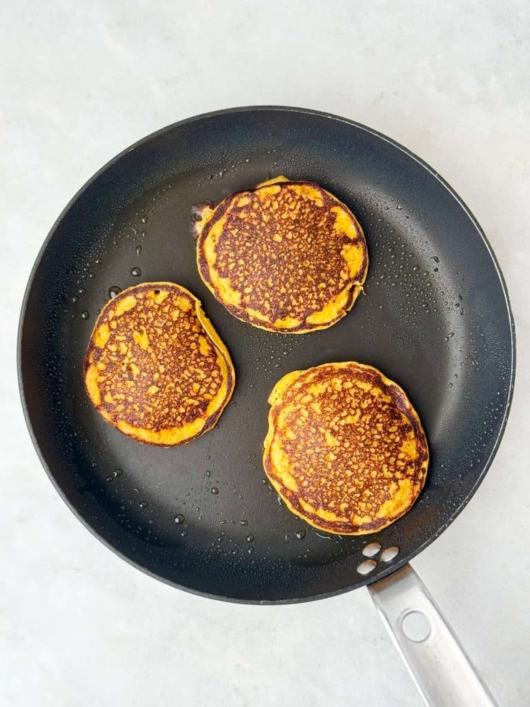Three pumpkin spice protein pancakes cooking in a nonstick skillet until golden brown.