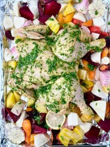Spatchcocked chicken coated in green herb rub placed over root vegetables ready to roast in the oven.