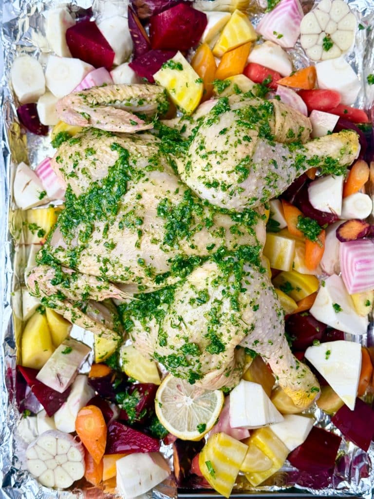 Spatchcocked chicken coated in green herb rub placed over root vegetables ready to roast in the oven.