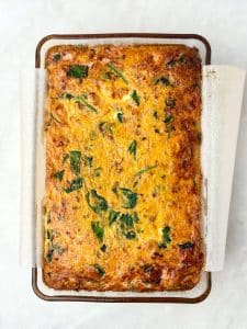 Golden-brown dairy-free breakfast casserole with sausage, shredded sweet potatoes, and spinach baked in a glass dish lined with parchment paper.