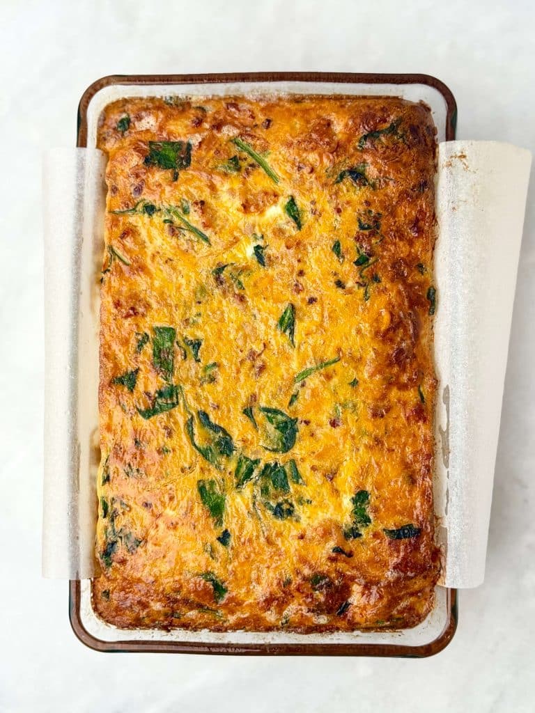 Golden-brown dairy-free breakfast casserole with sausage, shredded sweet potatoes, and spinach baked in a glass dish lined with parchment paper.