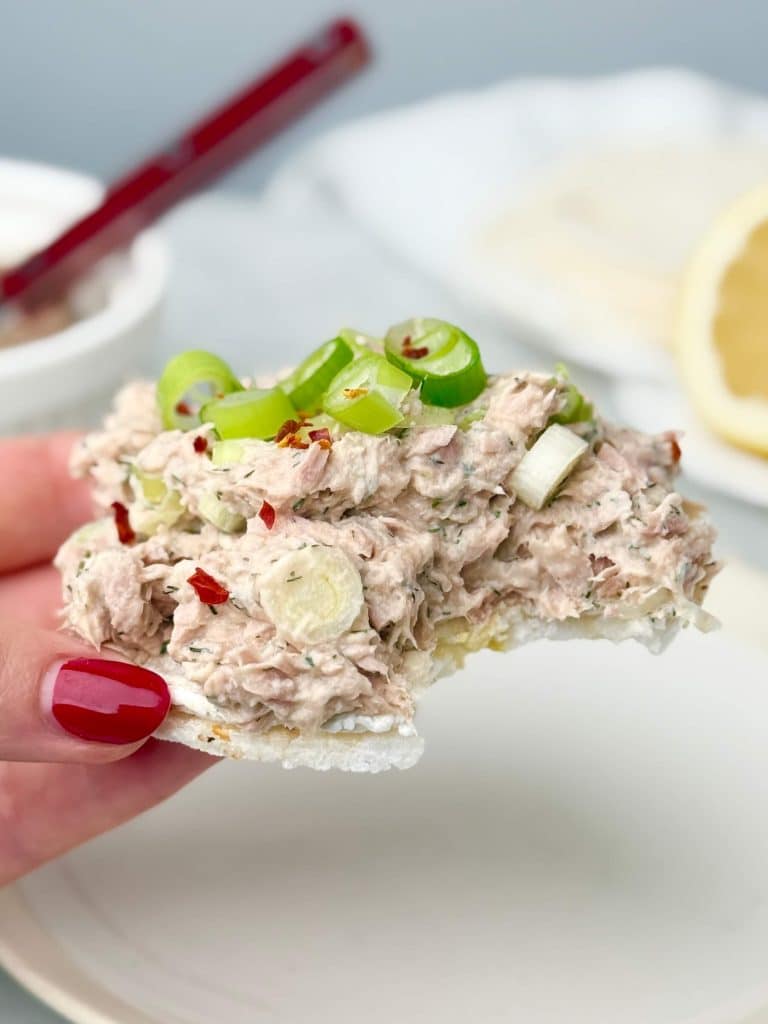 Close-up of a rice cake topped with creamy tuna salad, sliced green onions, and chili flakes, held in hand with a blurred lemon and bowl in the background.