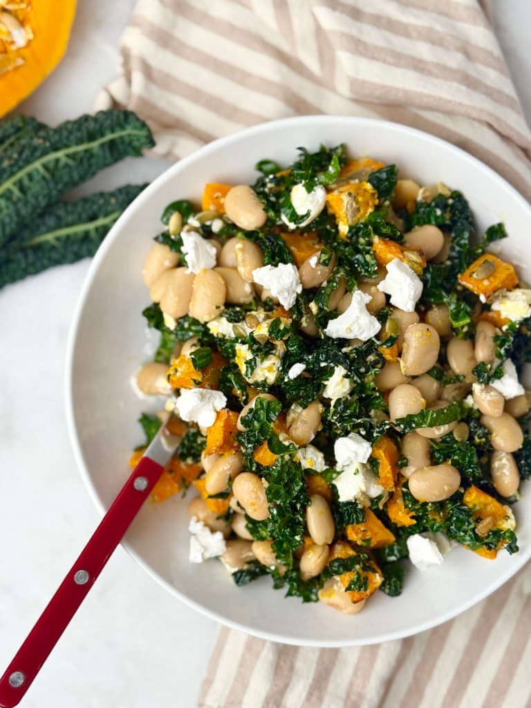 Overhead view of warm butter bean salad with roasted pumpkin, kale, and goat cheese in a white bowl on a striped cloth.