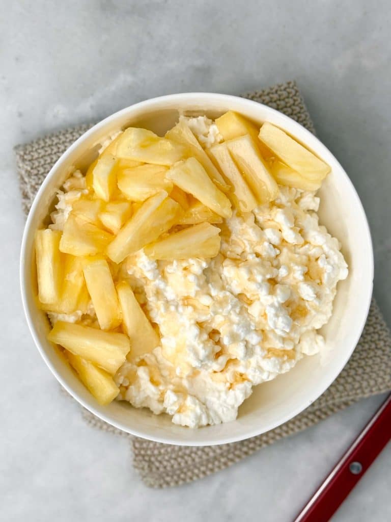 Bowl of low-fat cottage cheese topped with fresh pineapple chunks and a drizzle of honey, served on a light gray surface for a simple high-protein breakfast.