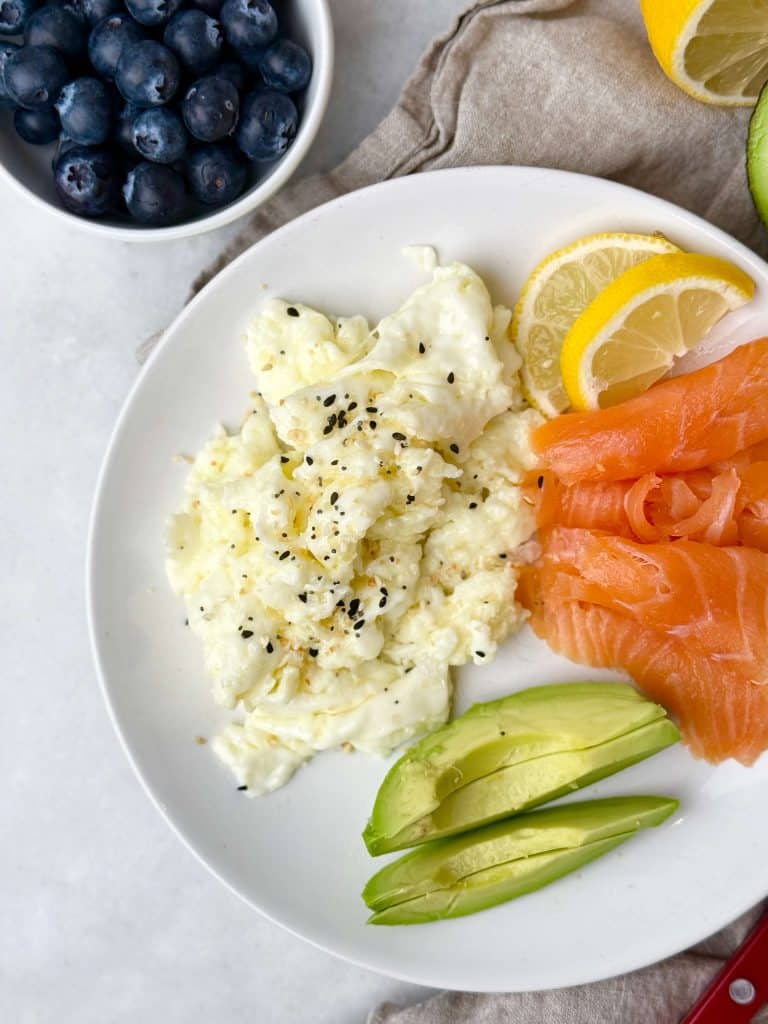 Plate of scrambled egg whites served with smoked salmon, sliced avocado, and lemon, with a bowl of fresh blueberries on the side for a light high-protein 300-calorie breakfast.