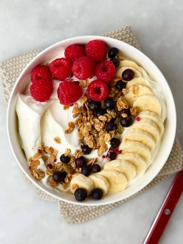 Creamy Greek yogurt bowl topped with sliced banana, raspberries, blueberries, and crunchy granola, photographed on a neutral background for a healthy high-protein breakfast.