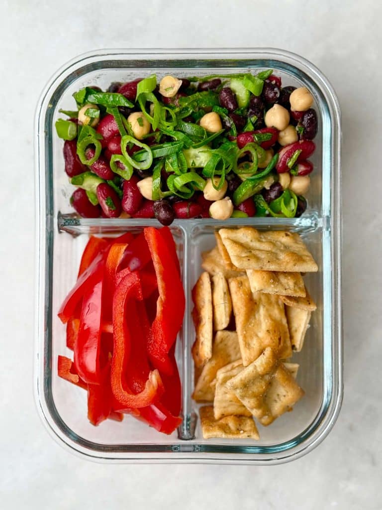 Three-compartment meal prep container with high-protein kidney bean salad, sliced red bell peppers, and whole grain crackers.