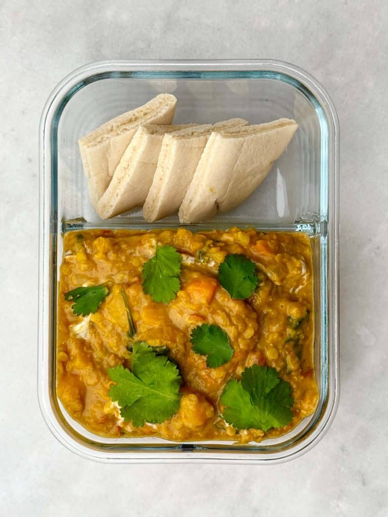 Glass container filled with creamy butternut squash red lentil curry topped with cilantro, served with sliced pita.