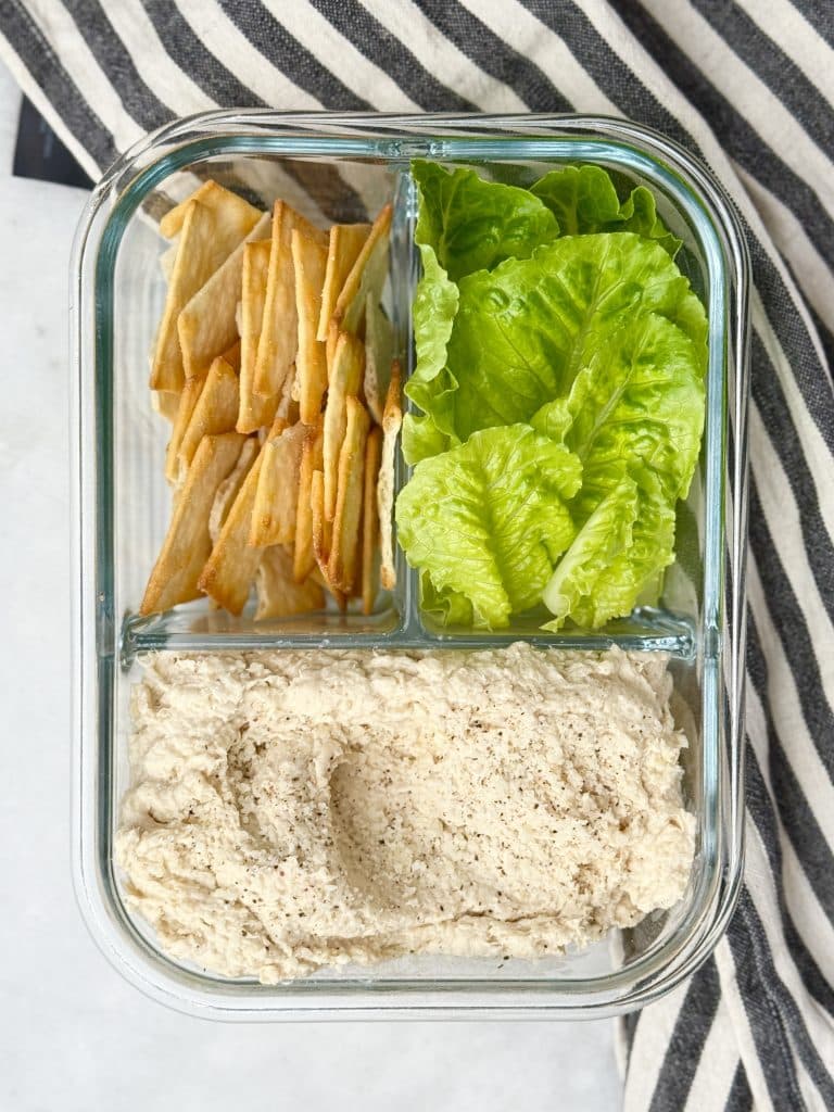 Meal prep container with creamy Caesar rotisserie chicken salad, lettuce leaves, and pita crackers for an easy high-protein lunch.