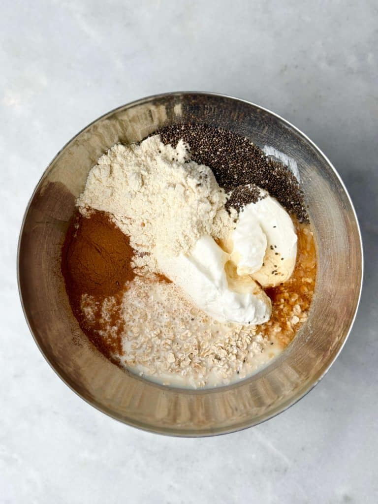 Overhead view of cinnamon roll overnight oats ingredients in a bowl, including rolled oats, cinnamon, chia seeds, Greek yogurt, protein powder, and almond milk before mixing.