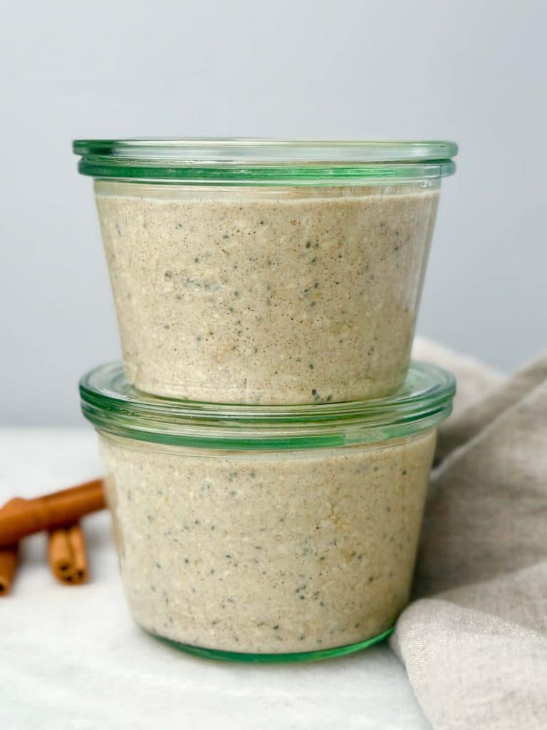 Two glass jars filled with creamy cinnamon roll overnight oats stacked on top of each other, with cinnamon sticks and a linen cloth in the background.