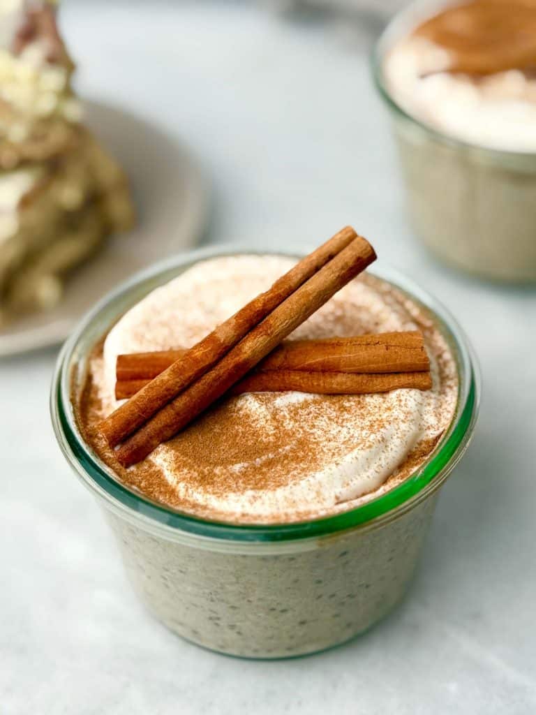 Glass jar of cinnamon roll overnight oats topped with a yogurt swirl, cinnamon dusting, and whole cinnamon sticks.