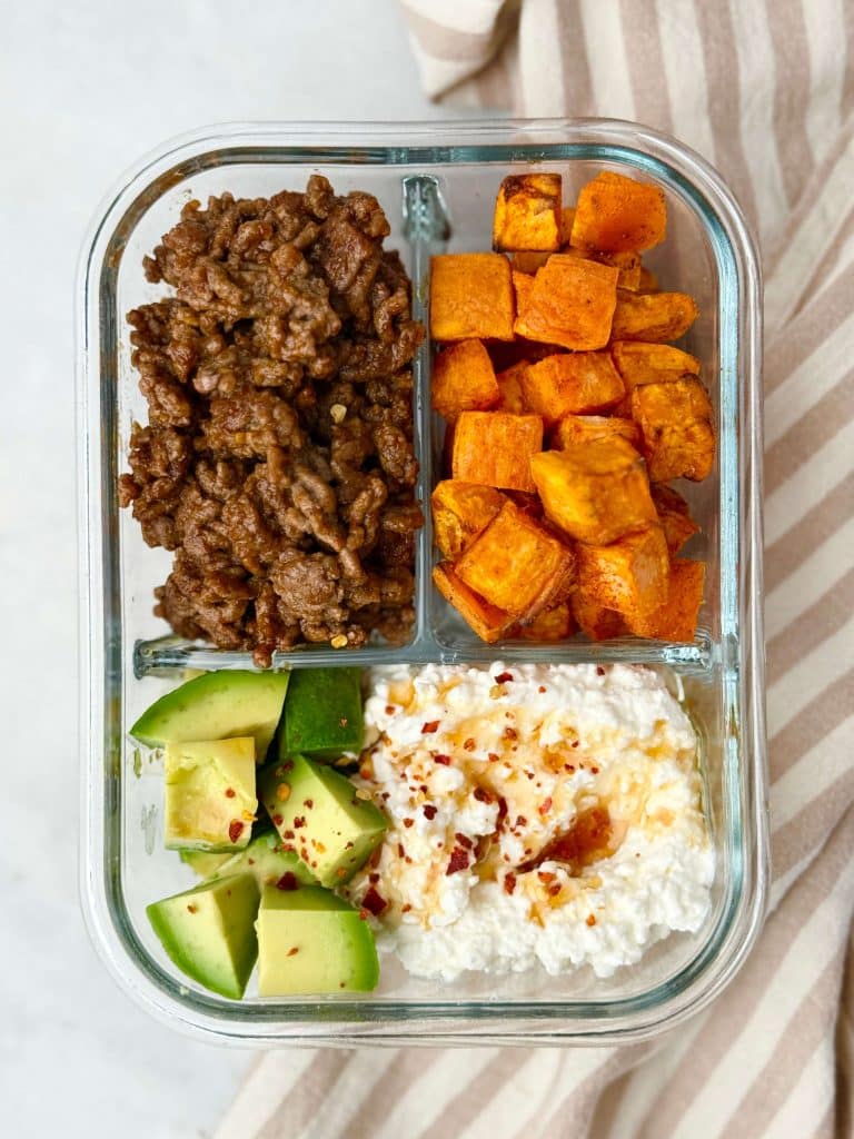 Glass meal prep container with cooked ground beef, roasted sweet potatoes, diced avocado, and cottage cheese drizzled with hot honey and chili flakes.