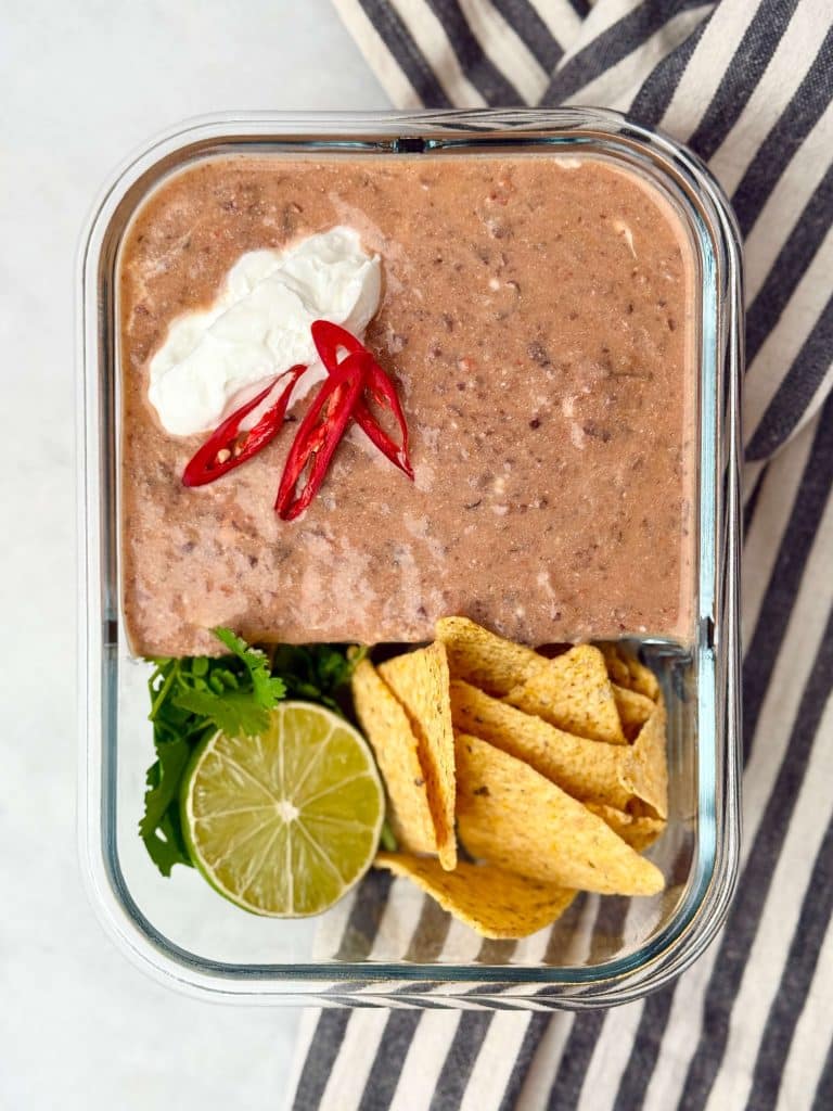 Meal prep container filled with creamy high-protein black bean soup topped with sour cream and sliced red chili, served with lime, cilantro, and tortilla chips.