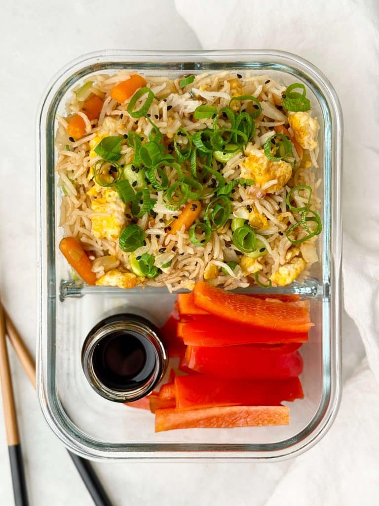 Glass meal prep container filled with high-protein egg fried rice topped with scallions, served with sliced red bell peppers and a small container of soy sauce.