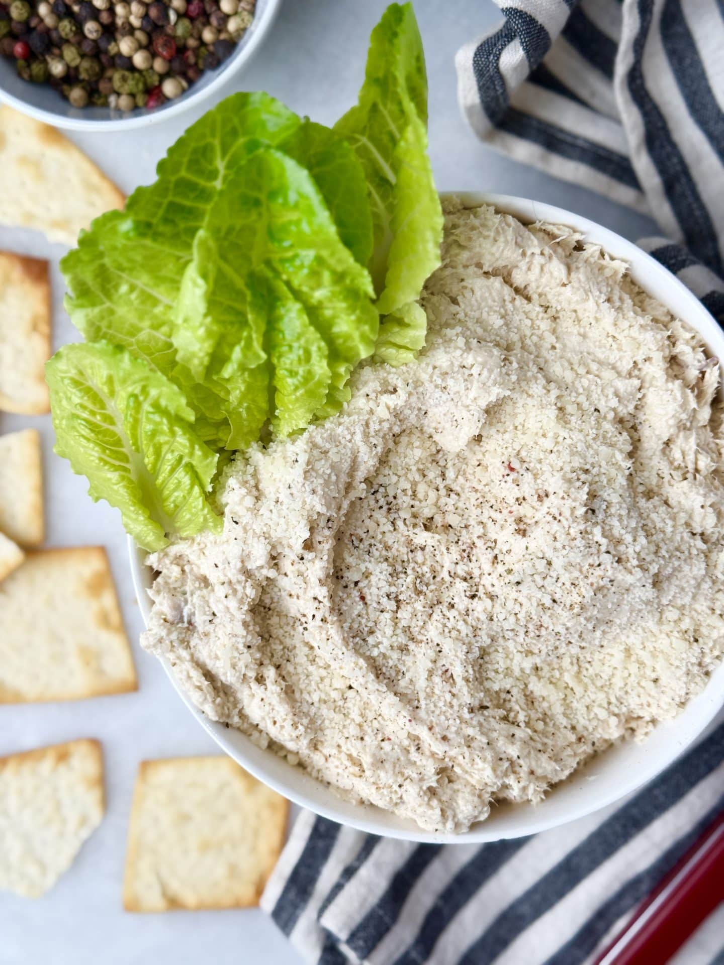 Rotisserie chicken salad with creamy Caesar dressing in a white bowl topped with Parmesan and romaine lettuce, surrounded by crackers.