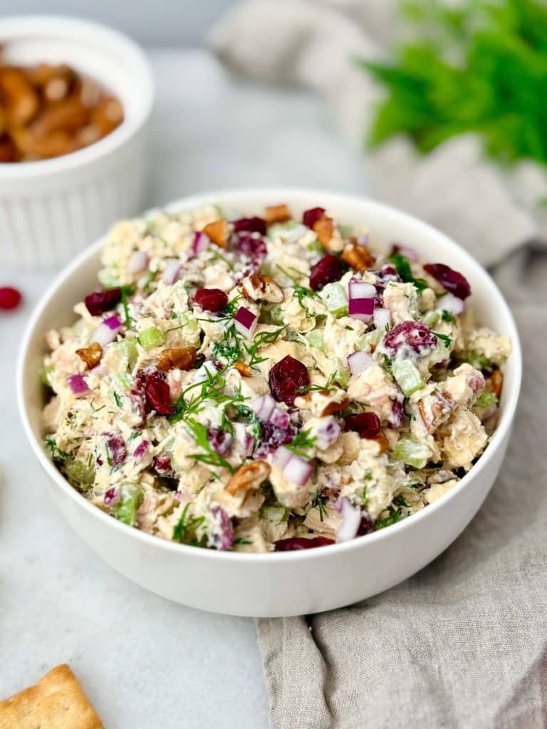 A side-angle view of creamy cranberry pecan chicken salad in a white bowl, showing the cranberries, pecans, celery, and fresh dill mixed throughout.