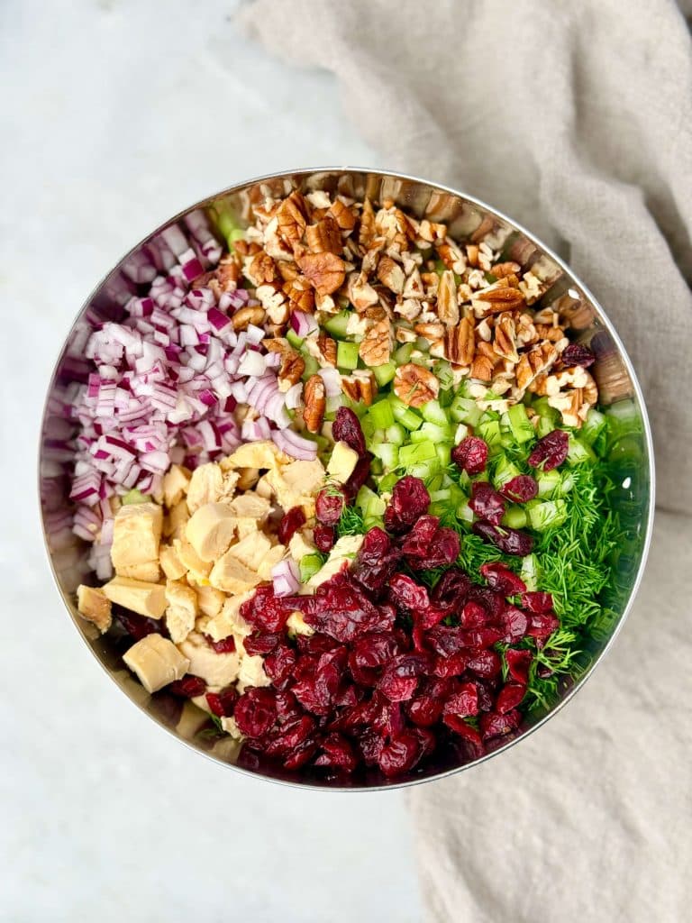 Ingredients for cranberry pecan chicken salad arranged in a bowl, including diced rotisserie chicken, dried cranberries, chopped pecans, celery, red onion, and fresh dill.