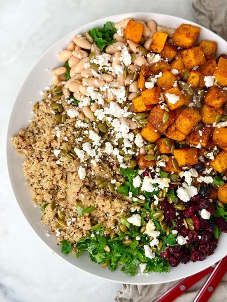 Harvest quinoa salad ingredients arranged in a bowl with kale, roasted squash, white beans, pumpkin seeds, dried cranberries, quinoa, and feta.