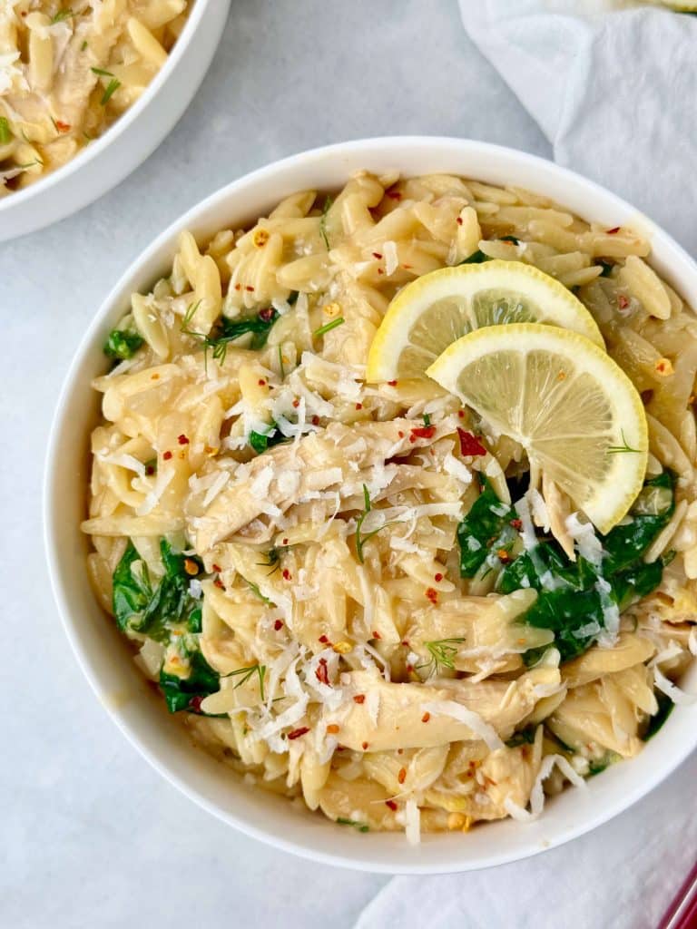 Creamy healthy lemon chicken orzo topped with parmesan, spinach, fresh dill, and lemon slices in a white bowl.