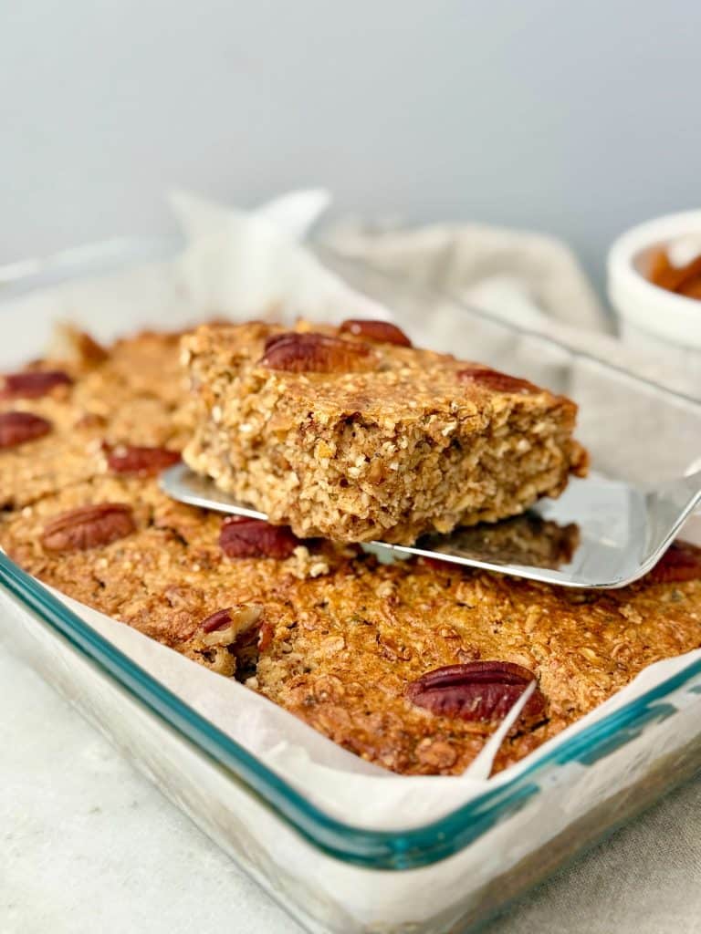 A warm square of pecan pie baked oatmeal being lifted from a glass baking dish, showing its soft, chewy texture and toasted pecan topping.
