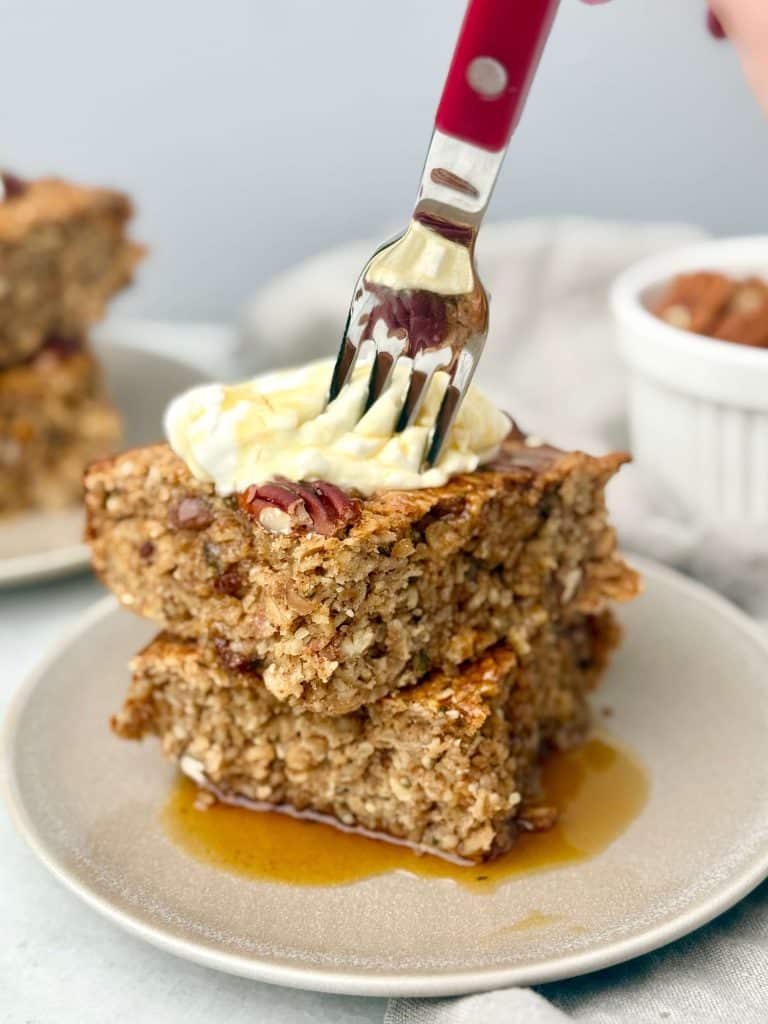 A fork breaking into a stack of pecan pie baked oatmeal topped with creamy yogurt and maple syrup, showing the tender, soft-baked texture.