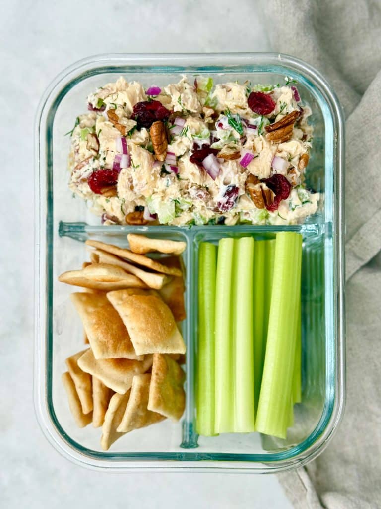 A meal prep container filled with cranberry pecan chicken salad, paired with celery sticks and crackers for an easy high protein lunch.