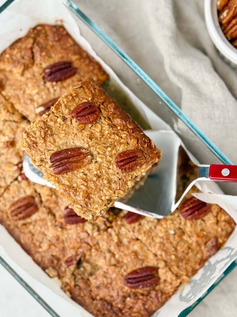 Overhead view of pecan pie baked oatmeal with one square lifted on a spatula, highlighting the golden edges and whole pecans on top.