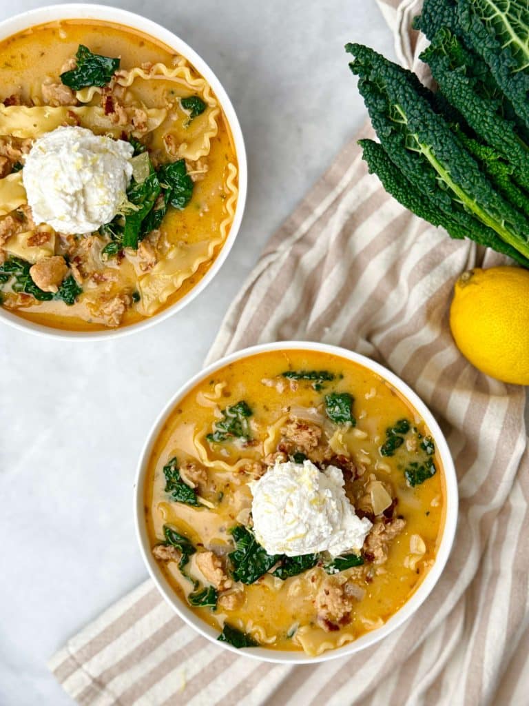 Two bowls of creamy white chicken lasagna soup with kale and lemon ricotta, shown with fresh lemons and striped linen.