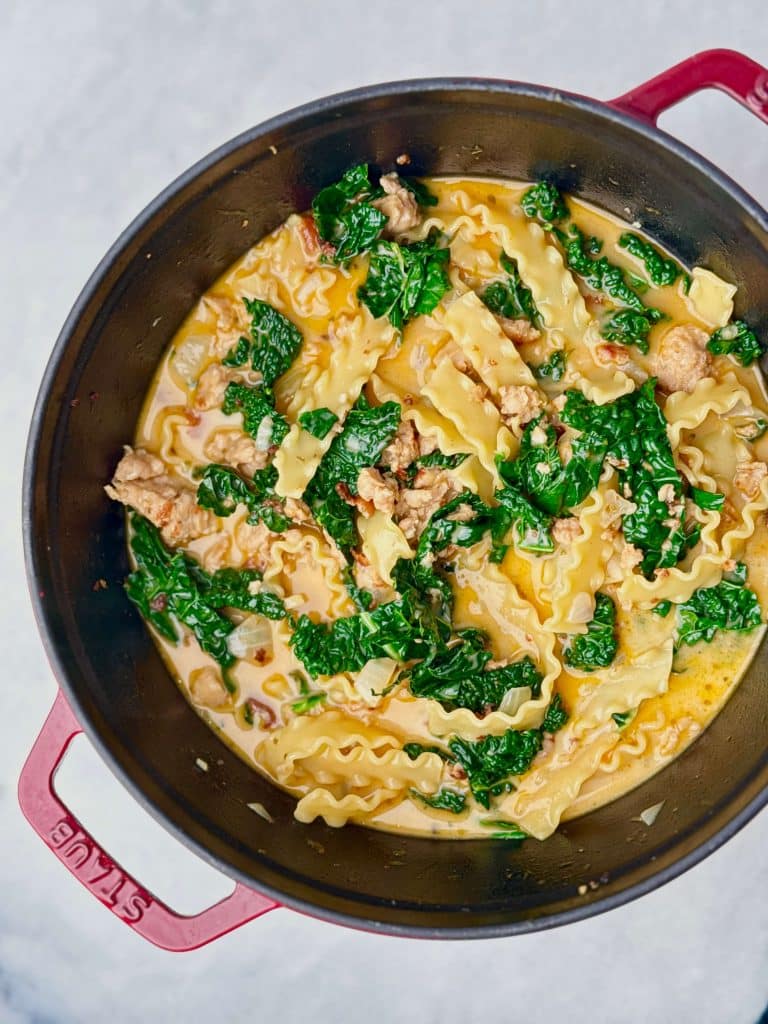 Top-down image of creamy white lasagna soup in a pot with chicken sausage, lasagna noodles, and fresh kale stirred in.