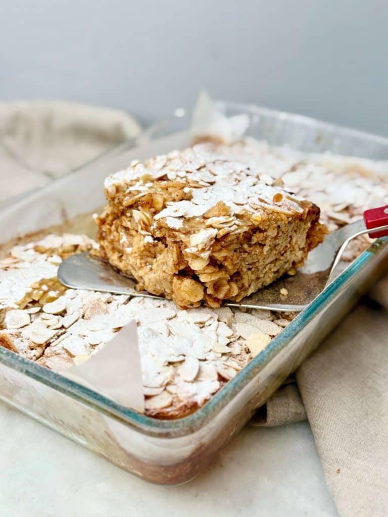 Almond croissant baked oatmeal square lifted from a glass baking dish, showing the soft texture and almond topping.