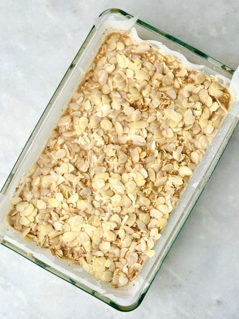 Almond croissant baked oatmeal before baking, topped with sliced almonds in a parchment-lined glass baking dish.