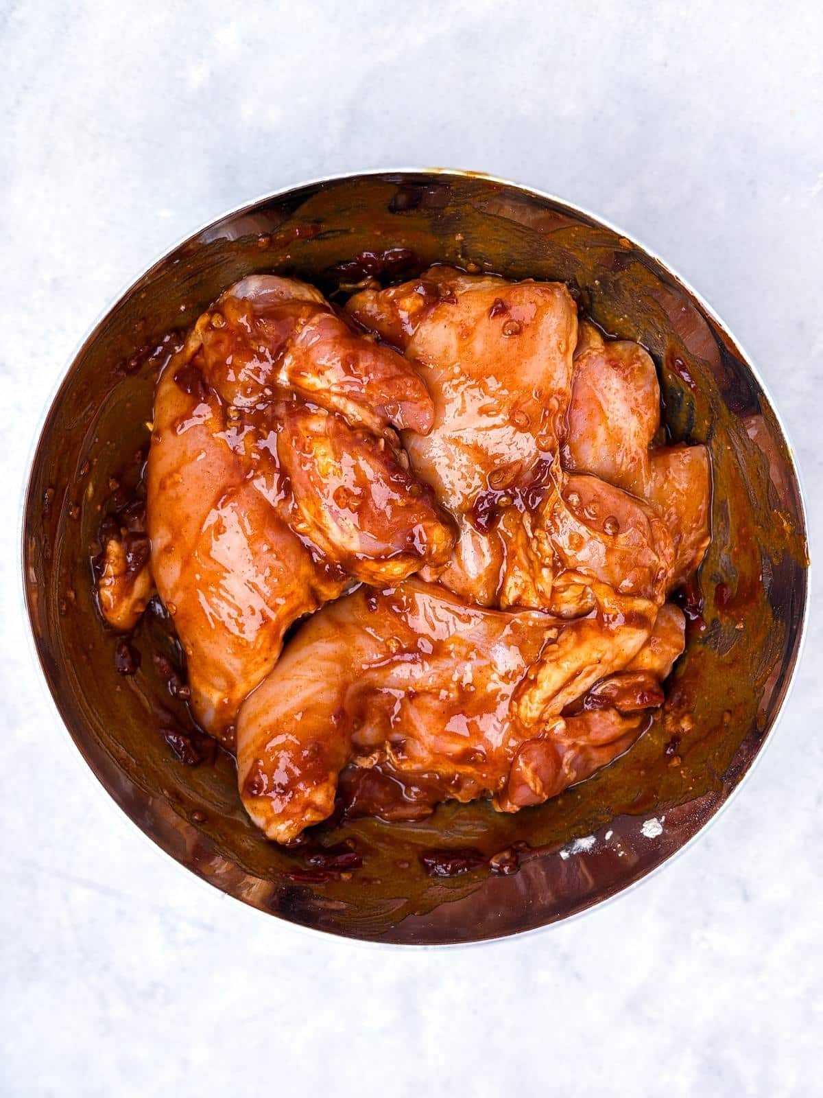 Chicken breasts tossed in BBQ marinade in a stainless steel mixing bowl