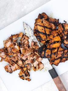 Grilled chicken thighs with char marks, half chopped on a white cutting board