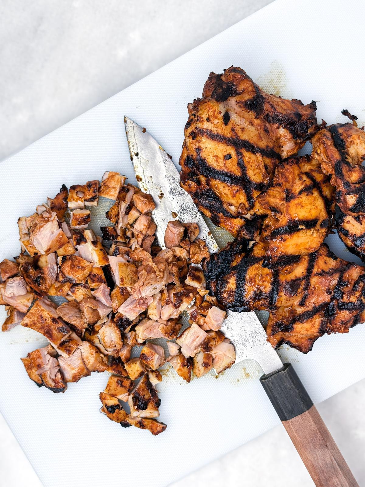 Grilled chicken thighs with char marks, half chopped on a white cutting board