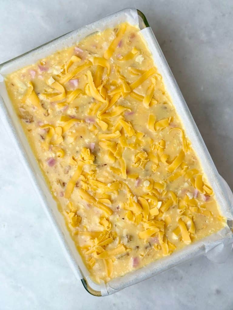 High protein egg casserole ready for meal prep, made with eggs, ham, shredded potato, and cheddar cheese in a baking dish before baking.
