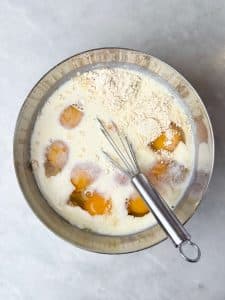 Egg mixture for a high protein egg bake made without cottage cheese, featuring eggs, milk, ham, onion, and shredded potato in a mixing bowl.