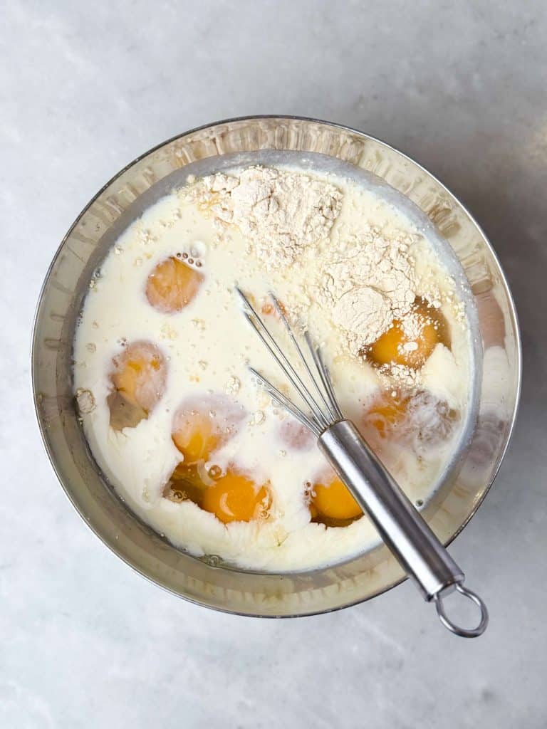 Egg mixture for a high protein egg bake made without cottage cheese, featuring eggs, milk, ham, onion, and shredded potato in a mixing bowl.