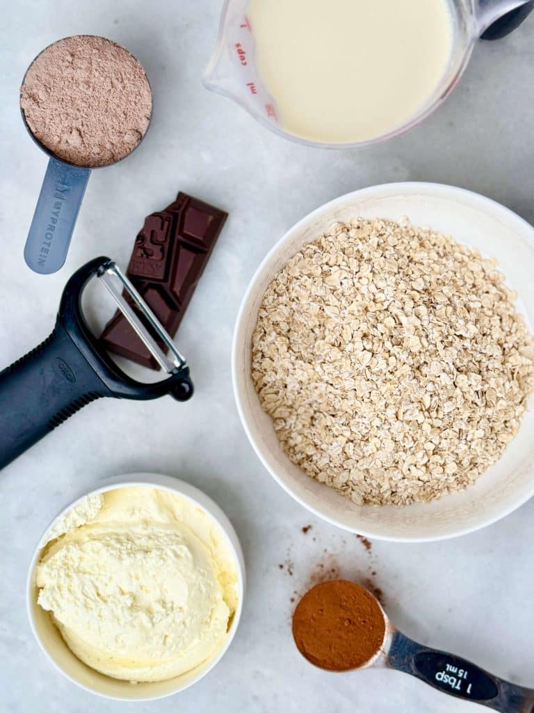 Ingredients for hot chocolate protein oats including rolled oats, cocoa powder, chocolate protein powder, milk, and whipped cream