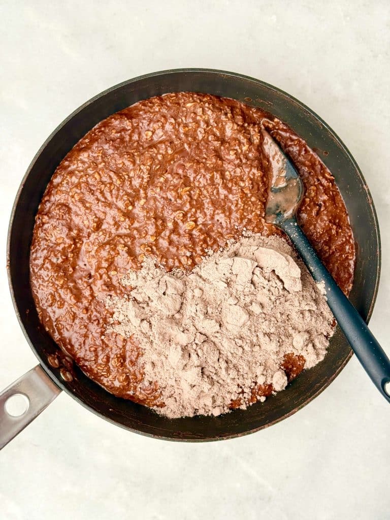 Adding chocolate protein powder to hot chocolate protein oats while cooking on the stovetop