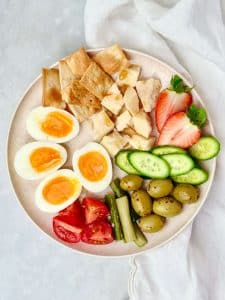 Healthy snack plate idea with soft-boiled eggs, cheese, olives, crackers, tomatoes, cucumbers, and strawberries.
