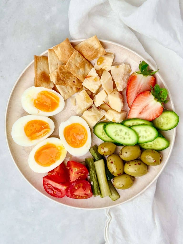 Healthy snack plate idea with soft-boiled eggs, cheese, olives, crackers, tomatoes, cucumbers, and strawberries.