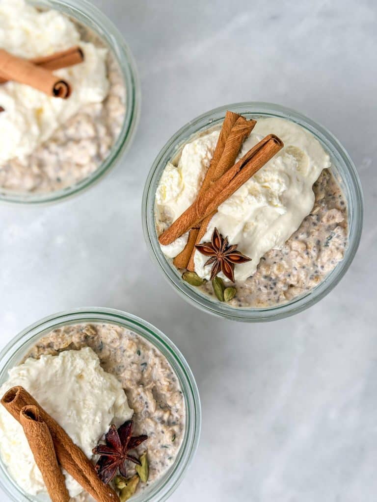 Overhead view of chai overnight oats topped with yogurt, cinnamon sticks, and star anise in meal prep jars.