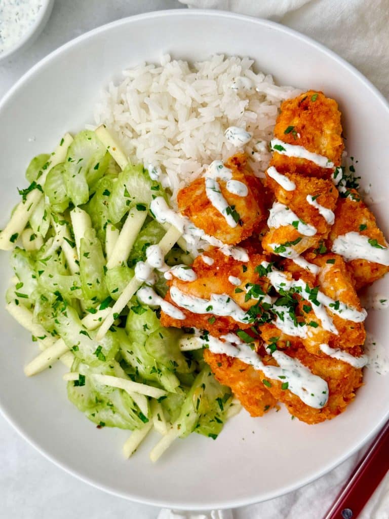 Air fryer buffalo chicken bites with celery apple slaw, white rice, and Greek yogurt ranch drizzle in a white bowl.