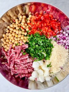 Ingredients for La Scala dense bean salad recipe including chickpeas, white beans, salami, mozzarella pearls, roasted red peppers, red onion, parsley, and grated parmesan.